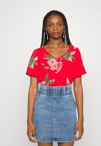 Blusa floral vermelha de mangas curtas, combinada com uma saia de denim azul claro com cintura elástica e bolsos.