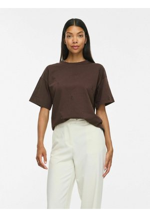Femme aux longs cheveux foncés portant une chemise ample à manches courtes marron foncé et un pantalon blanc taille haute, debout devant un fond uni.