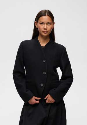 Femme aux longs cheveux raides portant un blazer noir boutonné et un pantalon noir sur un fond clair uni.