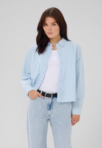 Camicia azzurro chiaro con maniche lunghe, in cotone, vestibilità relax. Indossata sopra una canottiera bianca, abbinata a jeans azzurro chiaro e una cintura nera.