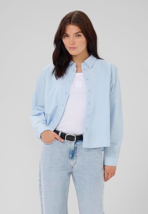 Light blue button-up shirt with long sleeves, cotton material, relaxed fit. Worn over a white tank top, paired with light blue jeans and a black belt.