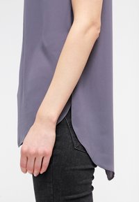 Blusa senza maniche di un leggero colore viola, realizzata in un tessuto morbido, con orlo curvo e fessure laterali, abbinata a jeans scuri.