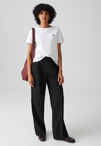 Weißes T-Shirt mit kleinem schwarzen Herzdesign, kombiniert mit lockeren schwarzen Hosen und einer maroon farbenen Schultertasche. Die Schuhe sind burgunderrot.