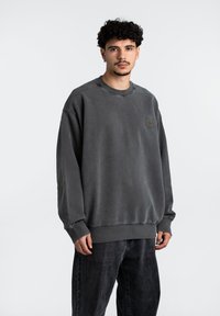 Sudadera gris de gran tamaño, hecha de una tela suave, con cuello redondo y puños acanalados, que presenta un pequeño parche con el logo en el pecho.