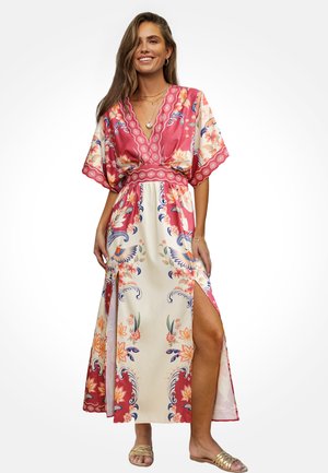 Vrouw die lacht, gekleed in een lange bloemenjurk met roze en crèmekleurige panelen, een zijsplit en gouden sandalen, tegen een witte achtergrond.