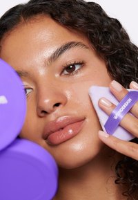 Contenitori di prodotti per la cura della pelle di colore viola circondano una persona che applica un applicatore di un leggero viola con un'etichetta etichettata "WONDERSKIN" sulla sua guancia.
