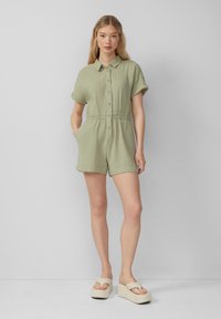Romper verde chiaro a maniche corte con abbottonatura frontale, colletto e vita elastica. Presenta due tasche laterali e una texture a coste. In mostra sandali con zeppa.