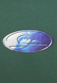 Logo ovale métallique avec un design stylisé de vague et de montagne sur un fond en tissu vert texturé.