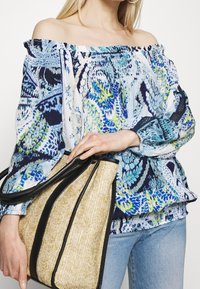 Off-shoulder bluse i blå og grøn med indviklede blomsterprint; letvægtigt stof; parret med en beige og sort tote taske.