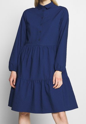 Woman wearing a long-sleeve, dark blue checkered dress with buttons down the front and a tiered, gathered skirt.