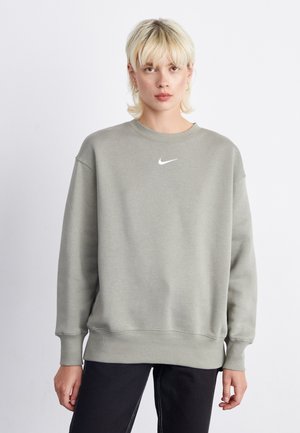 Sudadera gris hecha de una tela suave, con un cuello redondo y un pequeño logo blanco de Nike en el pecho. Pantalones negros visibles en la parte inferior.