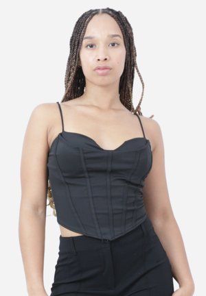 Femme aux longues tresses portant un corset noir ajusté à fines bretelles et un pantalon noir, debout devant un fond blanc uni.