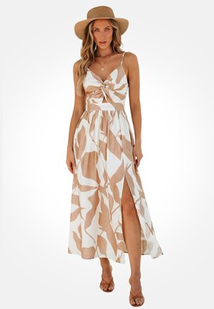Kobieta w sukience bez rękawów z beżowo-białym abstrakcyjnym nadrukiem, z rozcięciem z przodu, beżowych sandałach, warstwowych naszyjnikach i szerokim kapeluszu ze słomy.