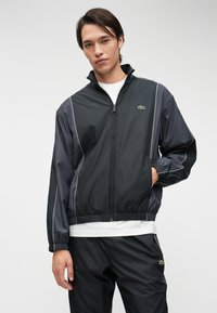 Μαύρο μπουφάν windbreaker με φερμουάρ μπροστά, ελαστικές μανσέτες, πλευρικές ραφές με λευκές λεπτομέρειες και ένα μικρό πράσινο λογότυπο στο στήθος.