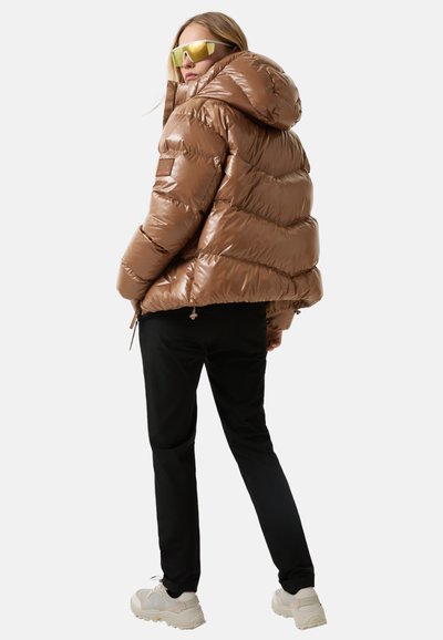 Person iført en brun, glansfuld pufferjakke med hætte, sorte bukser, kraftige hvide sneakers og store gultonede solbriller, som står med ryggen til.