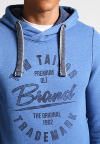 Blå hoodie gjord av mjukt material med dragsko i huvan, med en grafisk tryck i mörkgrått och kontrasterande innerfoder.