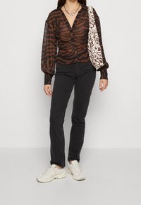 Blusa trasparente a righe zebra marrone e nera con scollatura a V, maniche a sbuffo lunghe e bottoni frontali, abbinata a jeans neri e sneaker chiare.