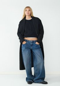 Zwarte lange jas over een zwarte cropped sweater, gecombineerd met hoge taille, wijde blauwe jeans. Zwarte ballerina's maken de outfit compleet.