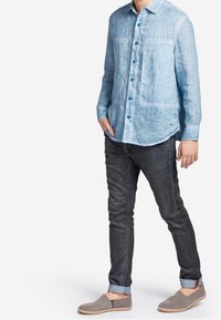 Camicia in lino azzurro chiaro a maniche lunghe con due tasche frontali e bottoni scuri, abbinata a jeans scuri e scarpe slip-on grigie.