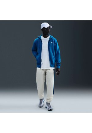 Mand med hvid kasket, blå lynlåshoodie, hvid T-shirt, cremefarvede joggingbukser og grå sneakers, stående mod en mørk gradient baggrund.