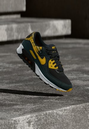 Μαύρο και κίτρινο αθλητικό παπούτσι Nike Air Max με λεπτομέρεια καρό, αιωρούμενο πάνω από γκρι σκάλες από σκυρόδεμα σε υπαίθριο χώρο.