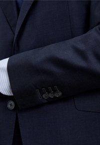 Manga de chaqueta de traje azul marino con tejido texturizado, tres botones oscuros y puño de camisa de vestir a rayas azul claro y blanco visible debajo.