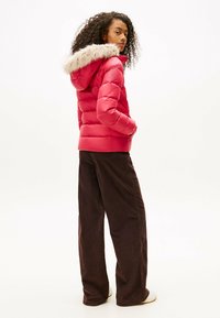 Czerwona kurtka puchowa z kapturem wykończonym futerkiem, z pikowanym wzorem i długimi rękawami, zestawiona z szerokimi spodniami w ciemnym brązie.