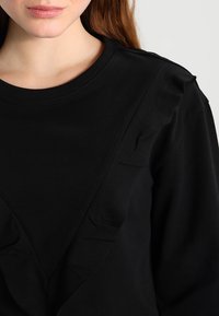 Nahaufnahme einer Person, die ein schwarzes Oberteil mit einer gerüschten V-förmigen Detail auf der Vorderseite und langen Ärmeln vor einem schlichten Hintergrund trägt.