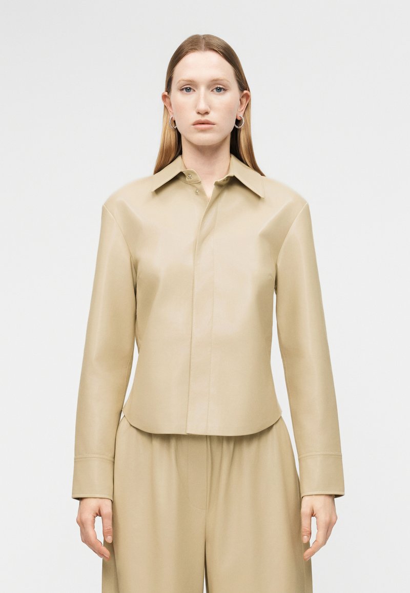 Žena s dlhými rovnými vlasmi, oblečená v béžovej blúzke s dlhými rukávmi a vo voľných nohaviciach, stojaca pred jednofarebným svetlým pozadím.