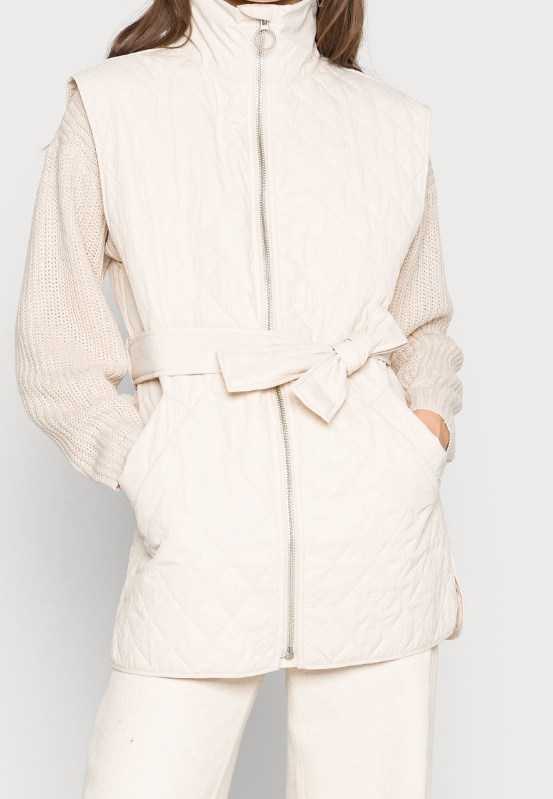 Gilet matelassé de couleur beige clair avec un col haut, une fermeture avant zippée et une taille ceinturée. Il comporte des poches latérales et des manches en tricot texturé.
