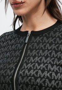 Jachetă neagră cu fermoar, având un model texturat de litere împletite. Materialul are un luciu subtil, cu guler striat și fermoar argintiu.