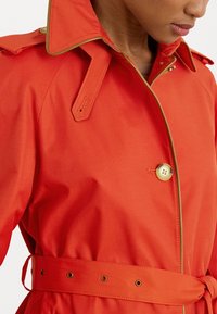 Heldere oranje trenchcoat met een brede kraag, gouden accenten en een riem in de taille. Voorzien van een knoopsluiting en een soepele stofstructuur.