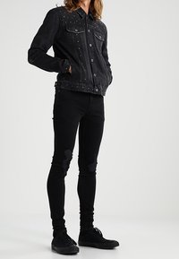 Chaqueta de mezclilla negra con detalles de tachuelas en los hombros, combinada con jeans ajustados negros con parches desgastados y zapatillas negras.