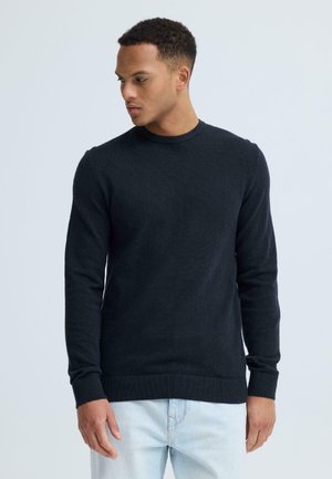 Man draagt een zwarte trui met lange mouwen en een ronde hals en een lichtblauwe spijkerbroek, kijkt naar links tegen een effen lichte achtergrond.