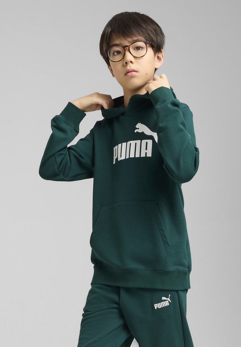 Zelena dukserica s kenguruvim džepom, sa velikim bijelim "PUMA" logotipom. Uparena sa usaglašenim zelenim trenirkama, oboje izrađeno od mekog materijala.