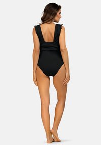 Kobieta stoi boso, odwrócona plecami, ubrana w czarny jednoczęściowy strój kąpielowy z niskim plecami i związanymi ramiączkami.