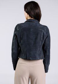Giacca di suede nera con un design aderente, dotata di un colletto a punta e dettagli delle cuciture, abbinata a pantaloni beige.
