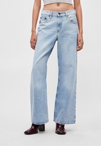 Φαρδύ τζιν ανοιχτού μπλε χρώματος με ψηλή μέση, κούμπωμα με κουμπιά και εμπρόσθιες τσέπες. Ομαλή υφή τζιν, ελαφρώς ξεθωριασμένο φινίρισμα, συνδυασμένο με σκούρα μποτάκια.