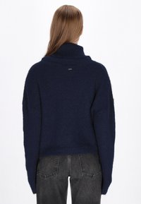 Vrouw met lang bruin haar in een donkerblauwe fuzzy trui en zwarte denim jeans, met de rug naar de camera gekeerd tegen een effen witte achtergrond.