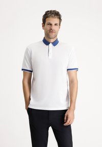 Camisa polo blanca con cuello y puños azules. Presenta una tapeta de tres botones, mangas cortas y una tela texturizada. Pantalones negros.