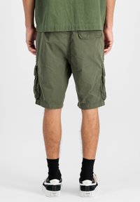 Shorts de tip cargo de culoare verde măsliniu, cu multiple buzunare laterale, margini răsucite și un material ușor, texturat. Poartă sosete negre și tenisi.