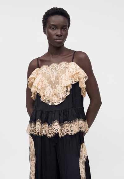 Γυναίκα που φοράει μαύρο αμάνικο φόρεμα με μπεζ δαντελένια φλοράλ βολάν στο στήθος, τη μέση και τις πλευρές των παντελονιών, στέκεται μπροστά σε απλό φόντο.