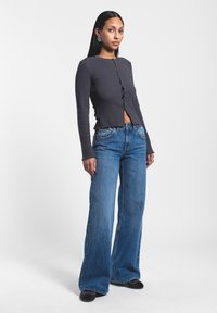 Top de manga larga de canalé gris oscuro con botón en el frente, combinado con jeans de pierna ancha azul claro y zapatos negros tipo slip-on. Pendientes plateados sencillos.