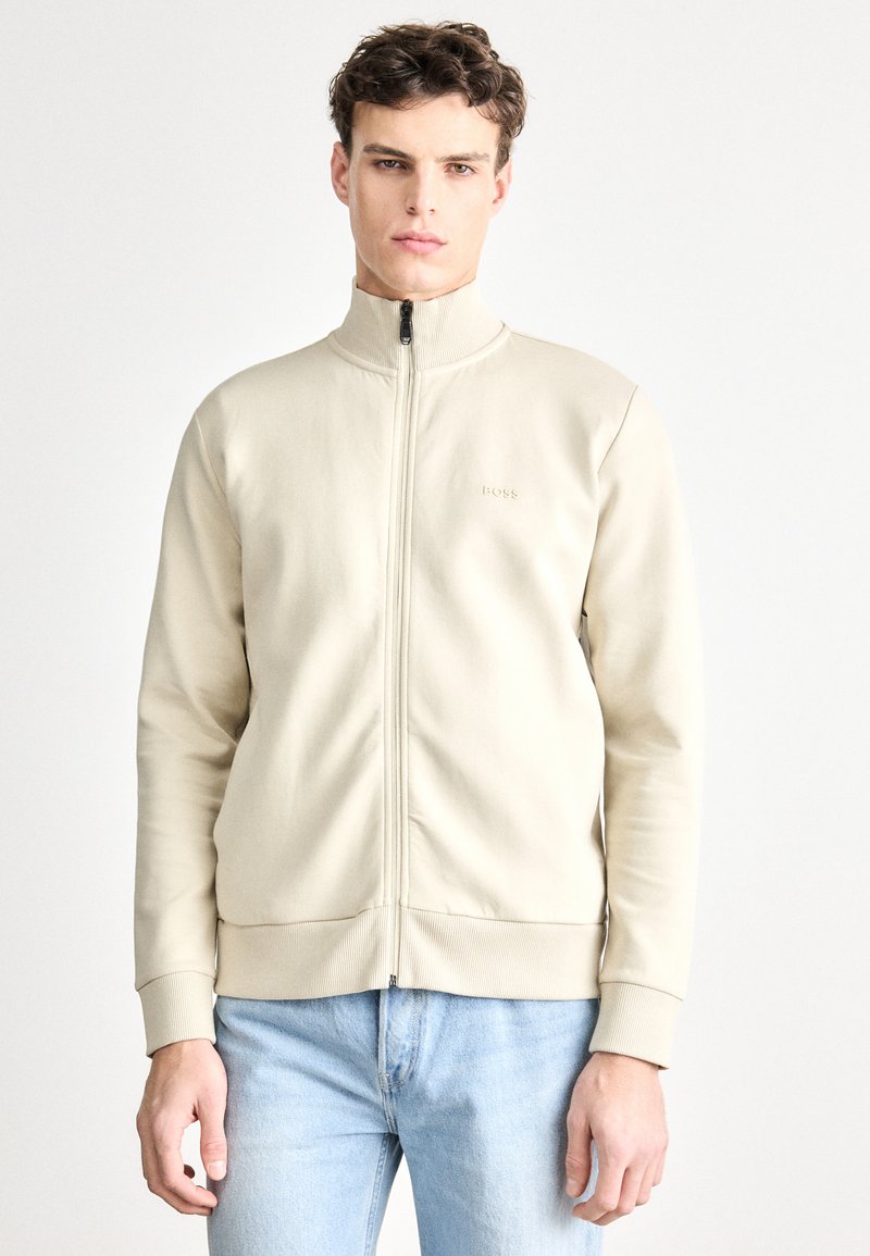 Bézs zipzáras dzseki magas gallérral, bordázott mandzsettákkal és derékrésszel; enyhén megjelenő márkajelzés a mellrészen. Világoskék farmerral viselve.