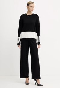 Maglione nero con striscia orizzontale bianca e dettaglio logo, abbinato a pantaloni neri a gamba larga. Tessuto a maglia morbida e vestibilità rilassata.