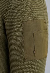 Olivgrüner Strickpullover mit einem strukturierten Muster, einem glatten Stoffpatch und einer Tasche am Ärmel, ergänzt durch dezente Nähdetails.
