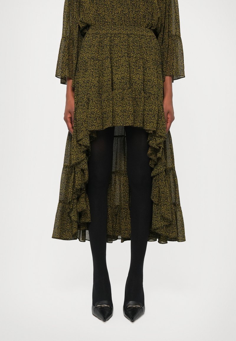Robe maxi à motifs verts avec des manches évasées et un ourlet festonné, portée sur des collants noirs et des escarpins noirs à bout pointu.