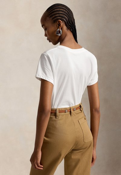 Femme aux cheveux tressés portant un t-shirt blanc, un pantalon marron, une ceinture marron et des boucles d'oreilles anneaux argentées, vue de dos.
