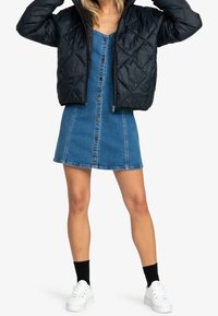 Veste matelassée noire à capuche portée sur une robe en denim bleu à boutons sur le devant. Portée avec des baskets blanches et des chaussettes noires.