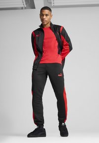 Mustmust ja punane sportlik spordikomplekt, mis sisaldab lukuga jope ja kitsenevaid pükse. Valmistatud siledast kangast, Puma logo aktsentidega ja kontrastsete paneelidega.